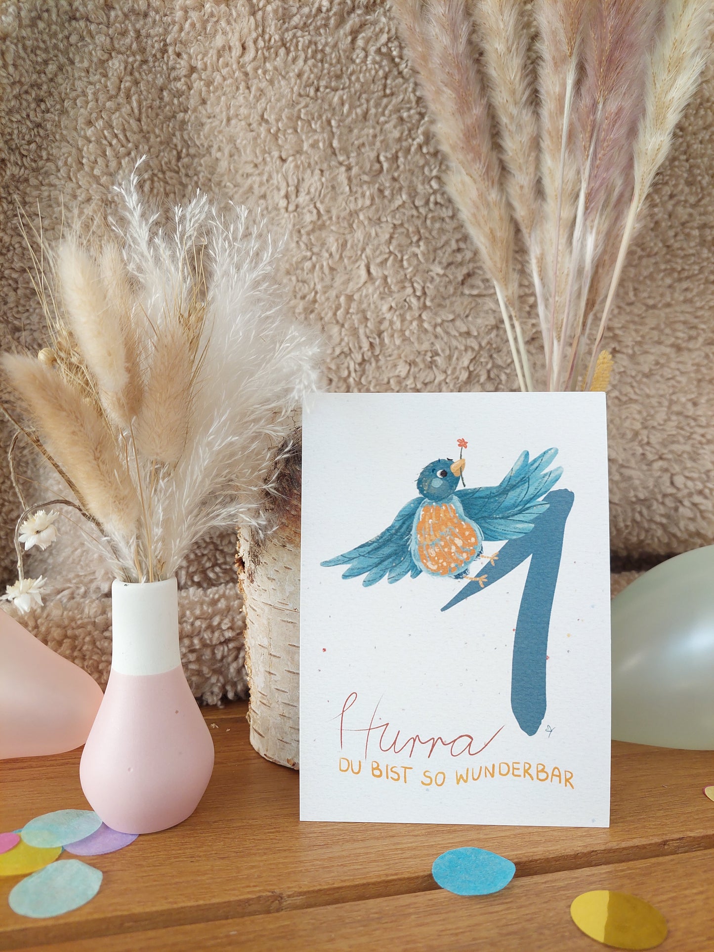 Erster Geburtstag -Postkarte. Blaue Eins mit blauem Vogel, der Blume im Schnabel hält. Konfetti im Hintergrund. Spruch: Hurra,du bist so wunderbar.