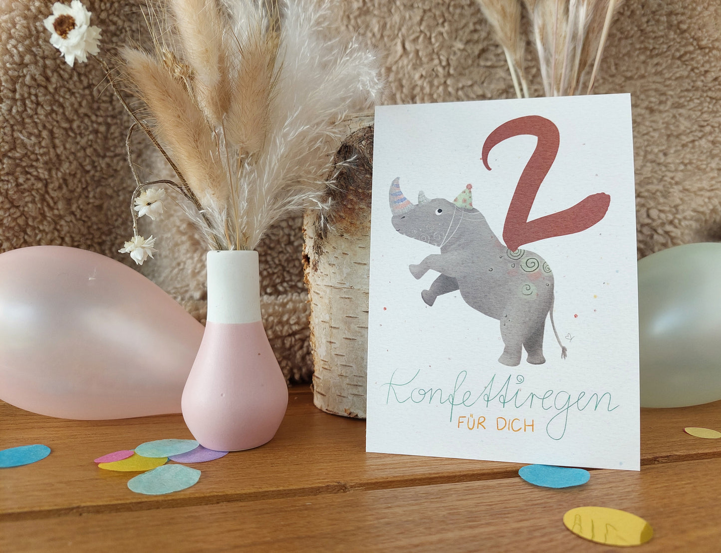 Postkarte zum zweiten Geburtstag. Buntes Nashorn mit Partyhut trägt große rote Zwei auf dem Rücken. Buntes Konfetti im Hintergrund. Spruch: Konfettiregen für dich