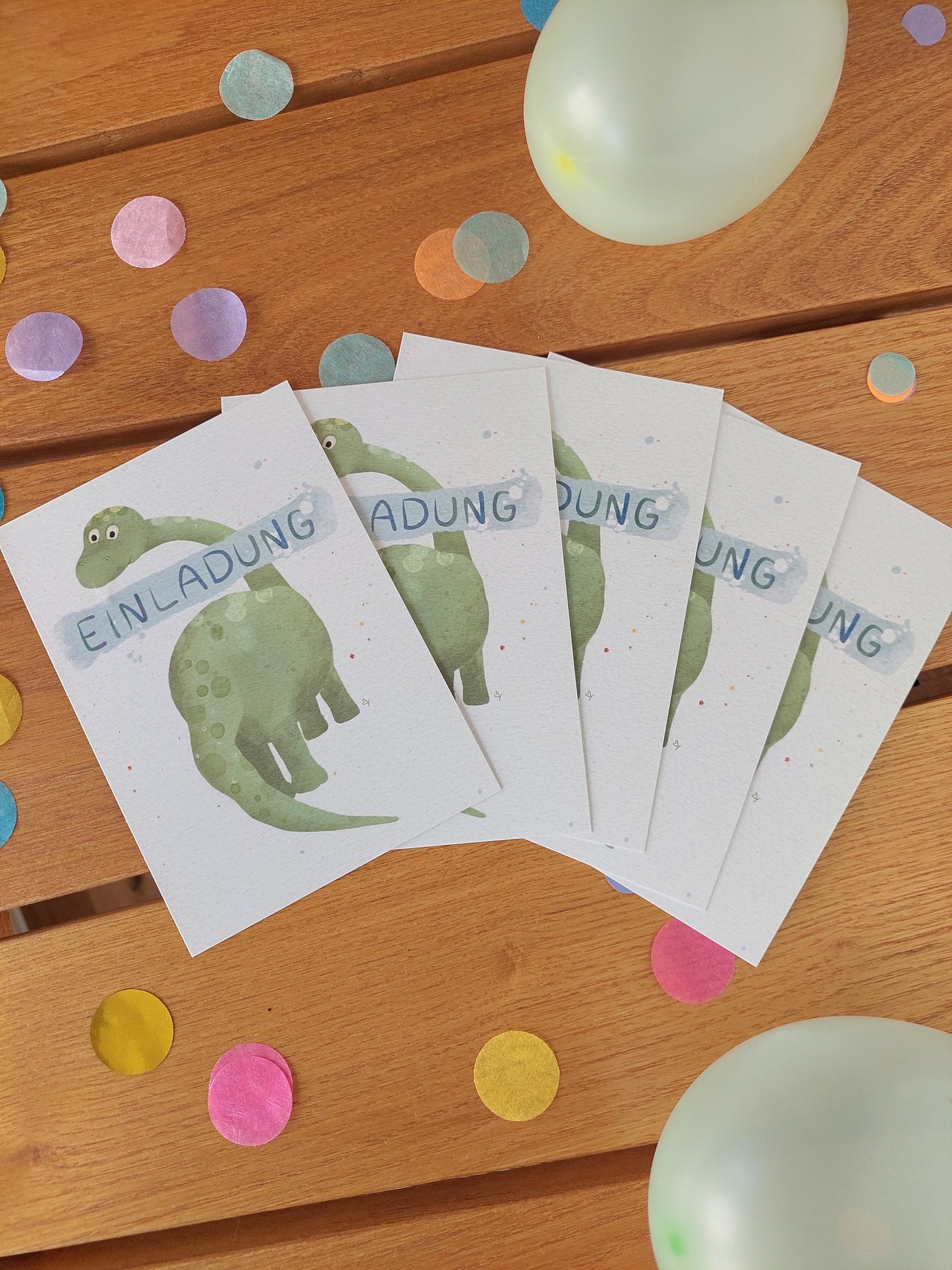 Fünf Dino-Einladungskarten im Set liegen zwischen buntem Konfetti und Luftballons auf einem Holztisch.