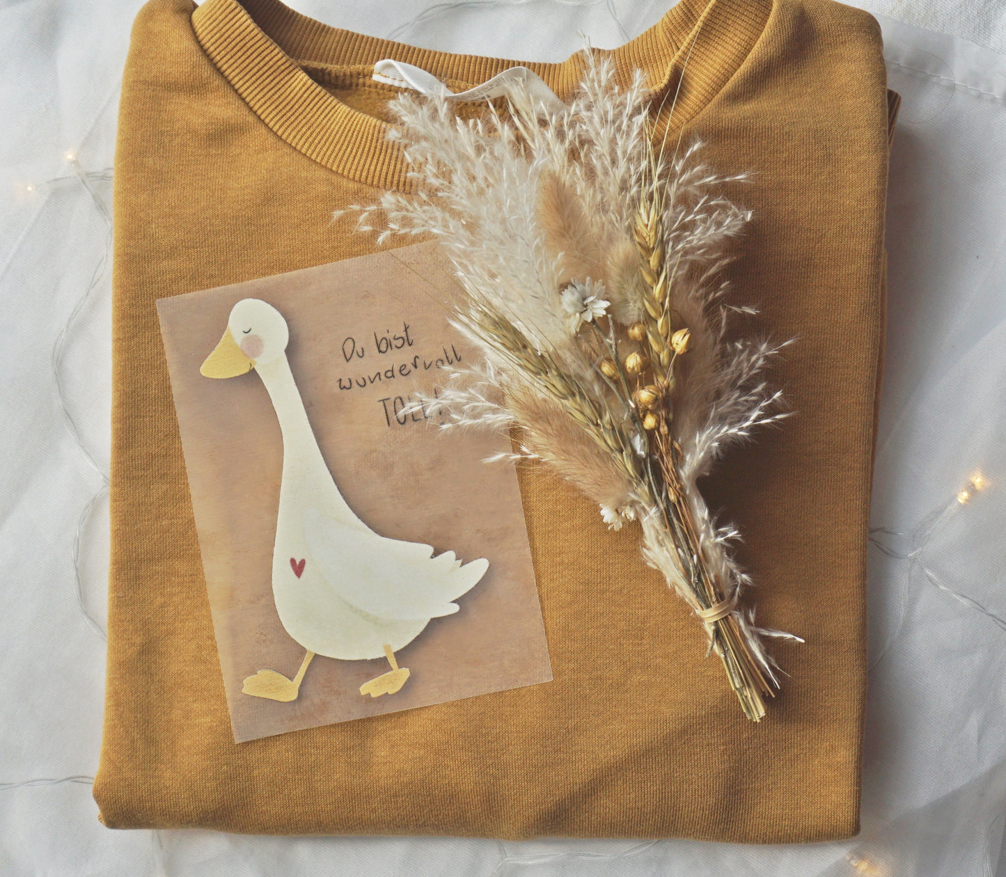 Bügelpatch von Gans mit Herz auf der Brust und Spruch: Du bist wundervoll toll! liegt auf einem senfgelben Pullover, miritime Zeichnung