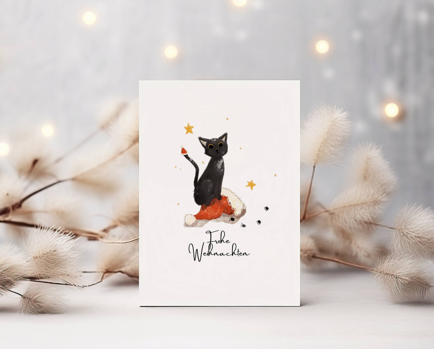 Grüße und Wünsche zu Weihnachten auf dieser süßen selbstgemalten Karte. Aquarell-Kreide-Zeichnung. Süße schwarze Katze mit Mondaugen sitzt auf Weihnachtsmannmütze. Kleine Mütze auch auf ihrer Schwanzspitze. Darunter Spruch: Frohe Weihnachten