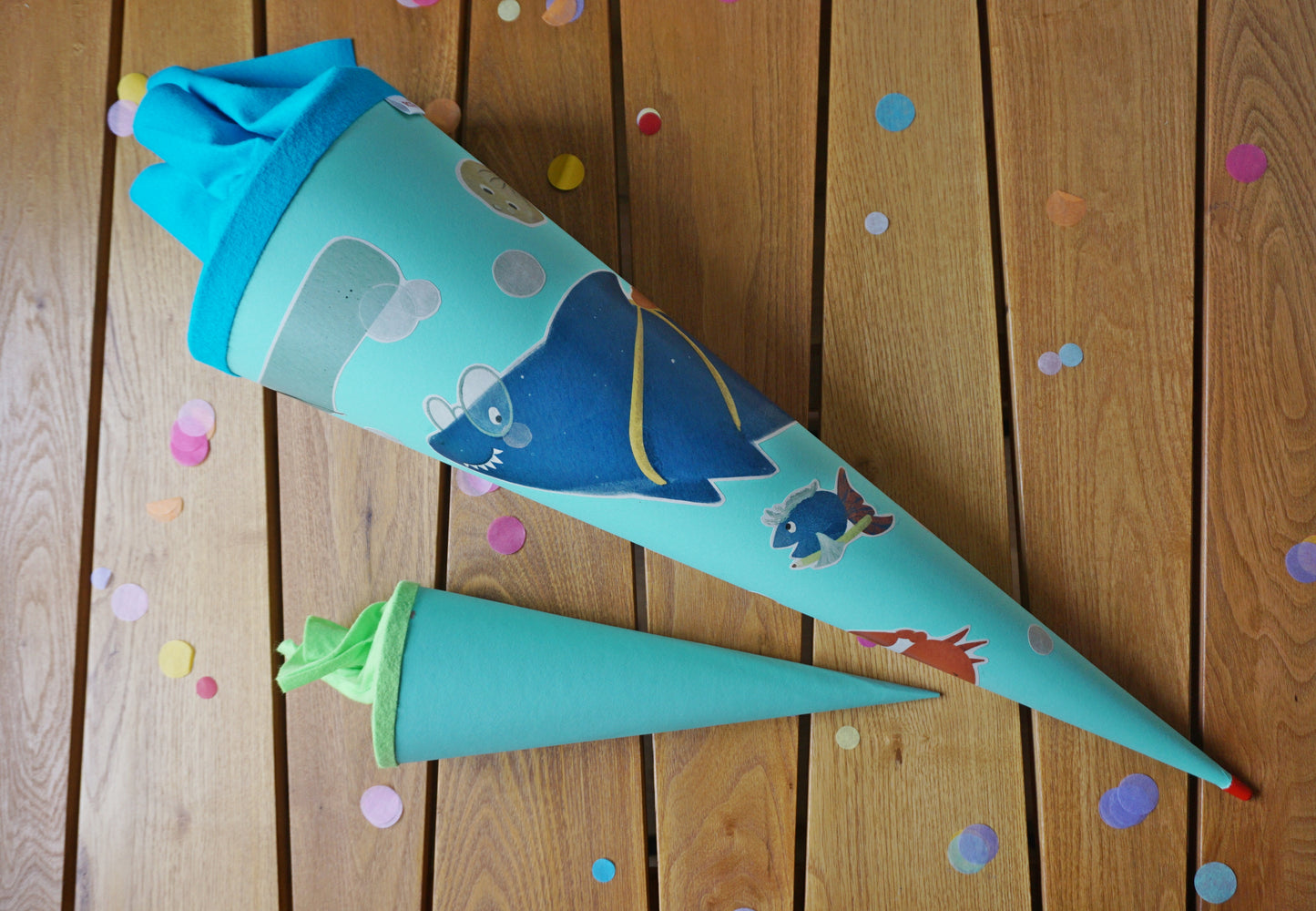 Schultüte selbst gestalten. Hier liegt eine türkise Schultüte mit strahlenden Meerestiere-Aufklebern auf einem Holztisch. Buntes Konfetti und eine kleine Geschwistertüte schmücken die Tisch.