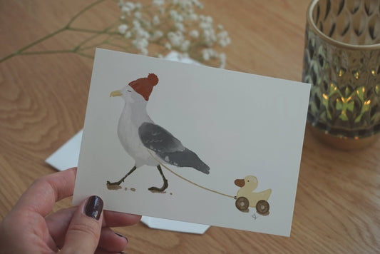 Das Bild Möwe spaziert mit Ente ist eine handgemalte Illustration. Man sieht die schöne, detaillierte Struktur des Motives. Eine entspannt blickende Möwe mit roter Mütze zieht eine gelbe Holzspielzeugente hinter sich her.