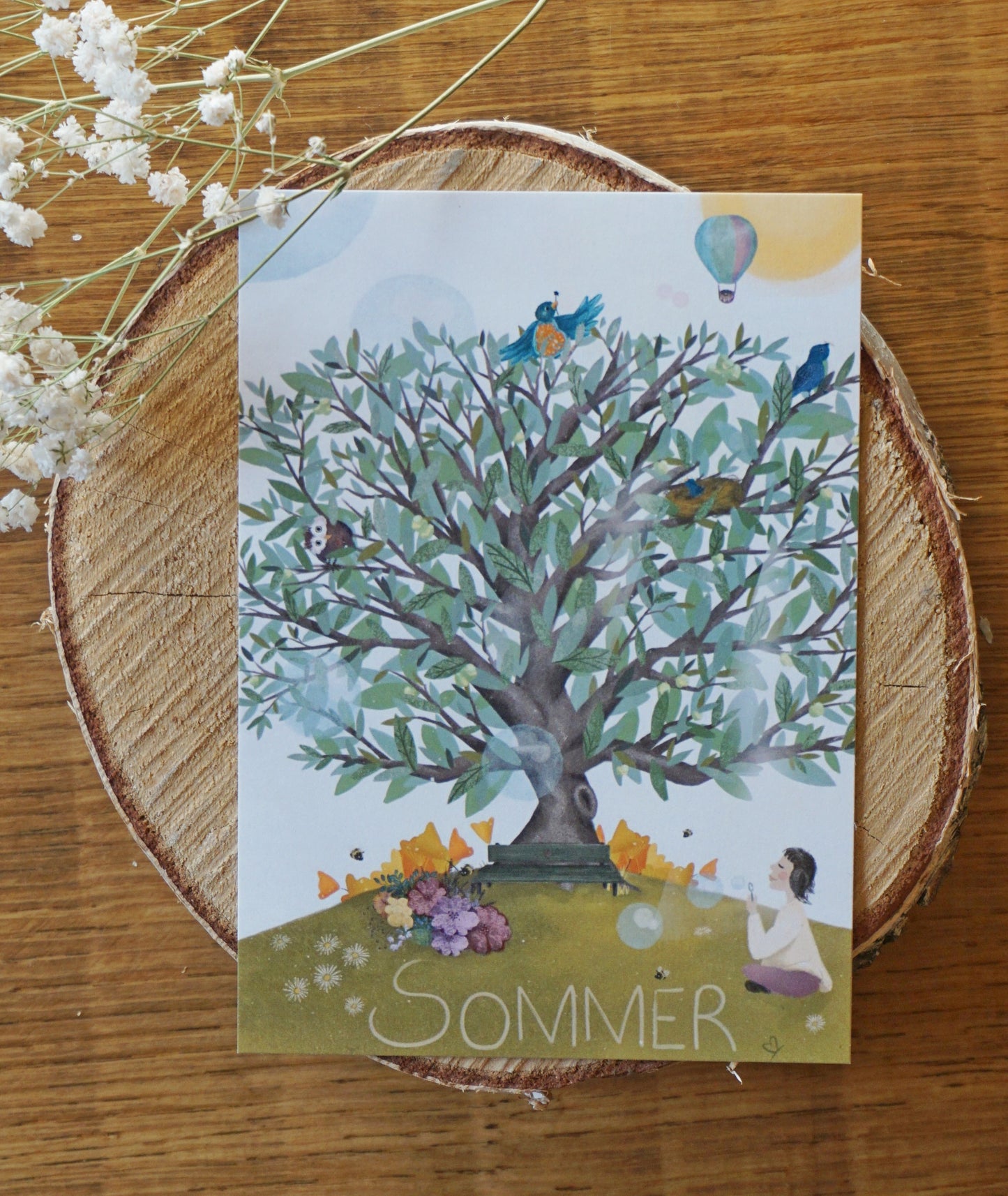 Baum mit grünen Blättern und grünen Äpfeln, Sonne und Heißluftballon im Hintergrund, auf der Wiese blühen Blumen, Bienen sammeln Nektar und ein Mädchen macht Seifenblasen, im Vogelnest sitzen kleine Voglekinder.